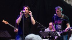 Bandmitglieder, zwei am Micro, dahinter ein Gitarrenhals und ein Schlagzeuger zu sehen