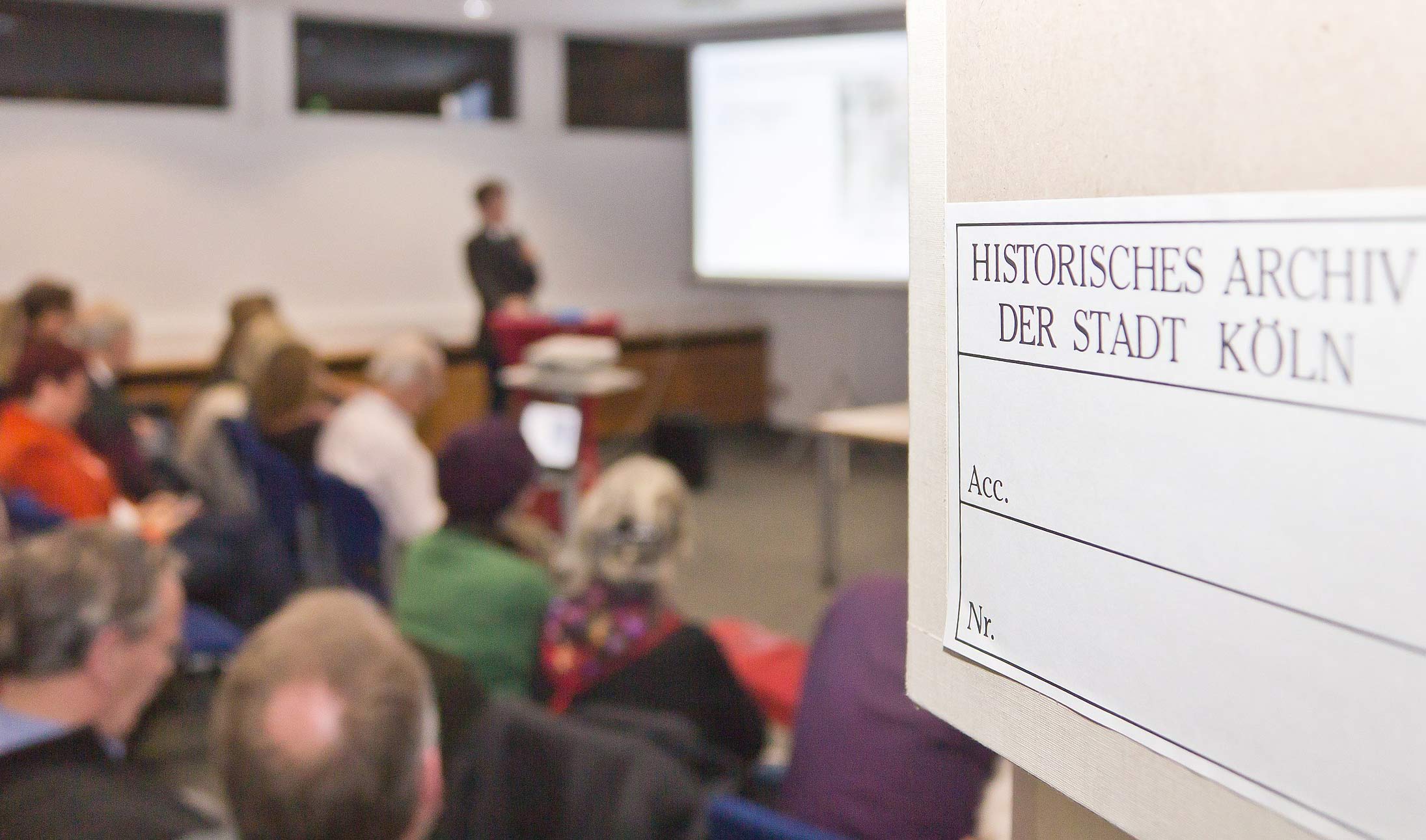 Menschen sitzen in Vortragsraum, im Hintergrund Vortragende und Leinwand (unscharf). Vorne rechts angeschnitten typischer Archivkarton mit Beschriftung Historisches Archiv der Stadt Köln