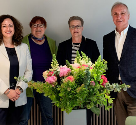Gruppenfoto hinter einem opulenten Blumenstrauß. Drei Frauen und ein Mann lächeln in die Kamera