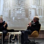 Zwei Männer sitzen auf der Bühne und schauen auf die Beamerwand, wo ein verscheneites Schwarzweißbld von Niedeckens Lebensmittelgeschäft zu sehen ist