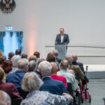 Vorsitzender am Rednerpult, Blick von hinten über das Publikum